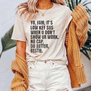 May include: A cream-colored t-shirt with black text that reads "YO, FAM, IT'S LOW KEY SUS WHEN U DON'T SHOW UR WORK. NO CAP. DO BETTER, BESTIE." The person is wearing white shorts and a mustard-colored cardigan.