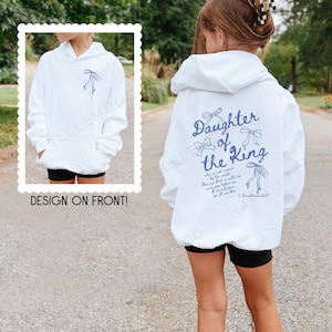 May include: White hoodie with a front pocket and a hood. The back of the hoodie features the text "Daughter of the King" in blue script, along with a bow graphic and a Bible verse. The front design is shown in a framed image.