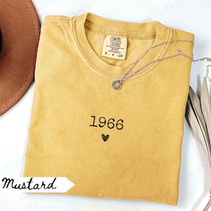 May include: A mustard yellow short-sleeve t-shirt with a crew neckline. The shirt features the embroidered black text 1966 above a small black heart symbol. The inner neck label reads Comfort Colors, 100% cotton, made in Honduras, size S.