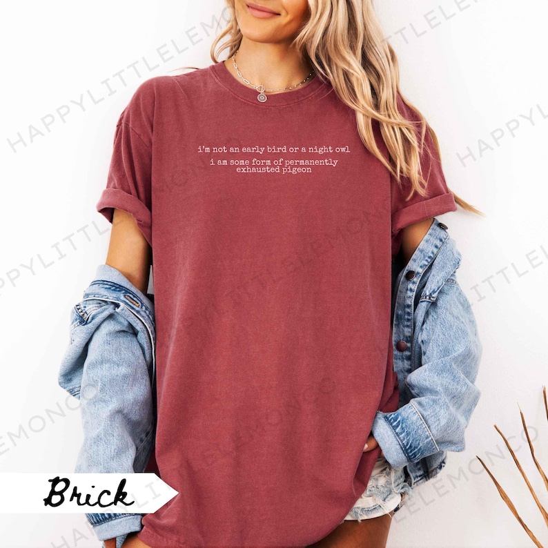 Puede incluir: Camiseta color ladrillo con mangas enrolladas. La camiseta presenta el texto blanco: "I'm not an early bird or a night owl, I am some form of permanently exhausted pigeon". Una chaqueta vaquera se lleva sobre la camiseta.