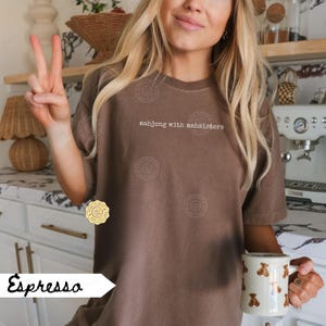 May include: Espresso-colored t-shirt with the text "mahjong with mahsisters" in white. The person is holding a white mug with brown bear illustrations. The person is giving a peace sign.