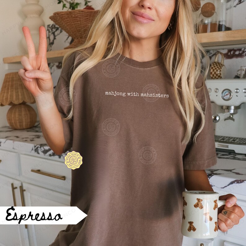 May include: Espresso-colored t-shirt with the text "mahjong with mahsisters" in white. The person is holding a white mug with brown bear illustrations. The person is giving a peace sign.