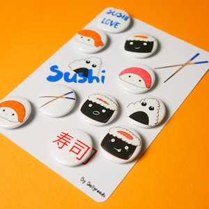 May include: A set of 10 round, colourful, printed buttons featuring various sushi designs. The buttons have white backgrounds and feature cute cartoon sushi characters with smiling faces. The text "Sushi Love" is printed on one button, and the text "Sushi" is printed on another button. The text "寿司" is printed on another button. The buttons are arranged on a white card with the text "By Dailygreets" printed at the bottom.