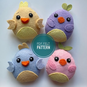 Puede incluir: Cuatro adornos de pájaros de fieltro en diferentes colores. Los pájaros son amarillos, morados, azules y rosas. Cada pájaro tiene un pequeño pico, ojos y una pequeña corona en la cabeza. Los pájaros están sentados sobre una superficie blanca. El texto "PDF FELT PATTERN" está en un círculo turquesa en el centro de la imagen.