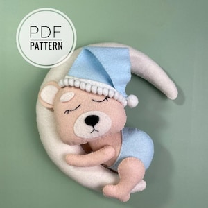 Puede incluir: Un oso de peluche rosa de fieltro con un gorro de dormir azul de fieltro está durmiendo en una luna creciente blanca de fieltro. La luna está unida a un fondo verde claro. El texto "PDF PATTERN" está en un círculo blanco en la parte superior izquierda de la imagen.