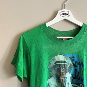 May include: Green t-shirt with a vintage image of Elton John wearing a white suit and sunglasses. The image is on a blue background with the text "Elton John" in white.