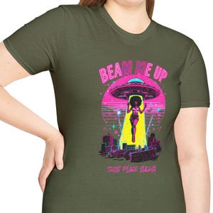 May include: Olive green t-shirt featuring a retro graphic of a UFO beaming up a woman. The design includes the text "BEAM ME UP" in pink and "THE PINK SEED" in a smaller font. The woman is wearing a yellow outfit.