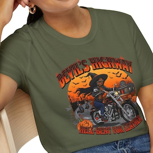 May include: Olive green t-shirt featuring a graphic of a witch riding a motorcycle on a highway, with the text "Devil's Highway" and "Hell-Bent for Leather". The design includes a full moon and pumpkins.