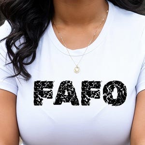 Può includere: T-shirt bianco con la scritta nera "FAFO" in un design strutturato. Sono visibili anche una collana d'oro e orecchini a cerchio dorati. La t-shirt è semplice.