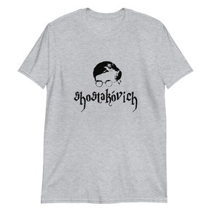 Può includere: Una t-shirt grigia melange con una silhouette nera di un uomo con gli occhiali e il testo "Shostakovich" in un font gotico.