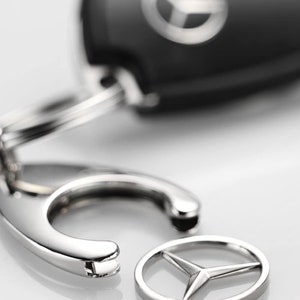 May include: A silver key chain with a black key fob featuring the Mercedes-Benz logo. The key chain has a silver ring and a silver hook.