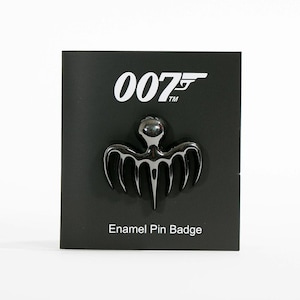 May include: A black enamel pin badge with a distinctive design, presented on a black card. The pin has a stylized, symmetrical shape with a central rounded element and multiple downward-curving lines. The card displays the "007" logo in white.