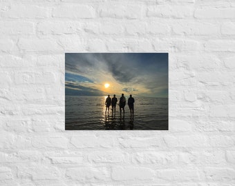 May include: Silhouettes of four people standing in shallow water, facing away from the camera, with the setting sun in the background. The sky is a mix of blue and gray with clouds.
