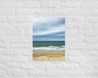 May include: A panoramic view of a sandy beach with blue ocean waves crashing on the shore. The sky is a light blue with white clouds.