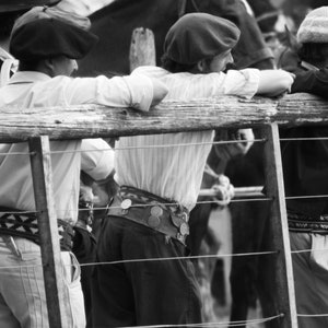 May include: Black and white photo of several people wearing traditional gaucho attire, including berets, button-down shirts, and decorative belts. They are leaning on a wooden fence, observing an event.
