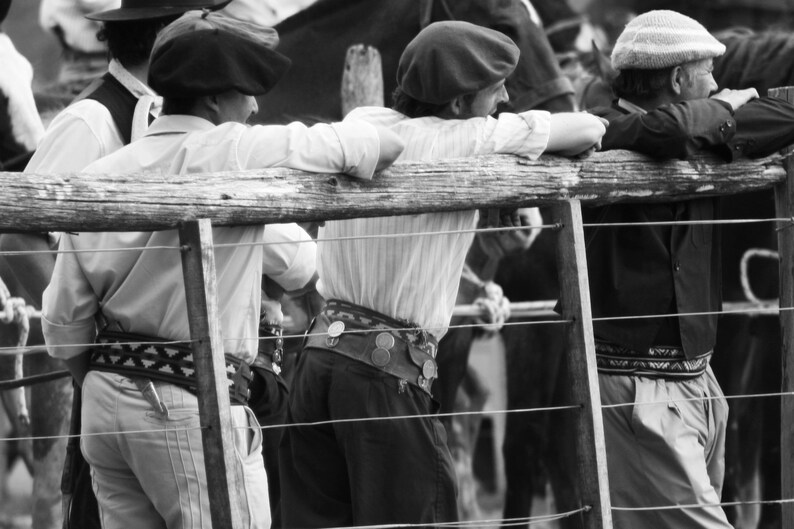May include: Black and white photo of several people wearing traditional gaucho attire, including berets, button-down shirts, and decorative belts. They are leaning on a wooden fence, observing an event.