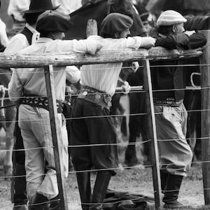 May include: Black and white photograph of several people leaning on a wooden fence, observing horses. The people are wearing traditional gaucho attire, including berets, button-down shirts, and riding boots. The scene evokes a sense of rural life.