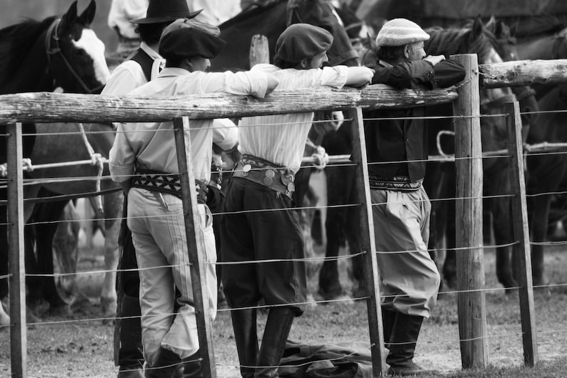 May include: Black and white photograph of several people leaning on a wooden fence, observing horses. The people are wearing traditional gaucho attire, including berets, button-down shirts, and riding boots. The scene evokes a sense of rural life.