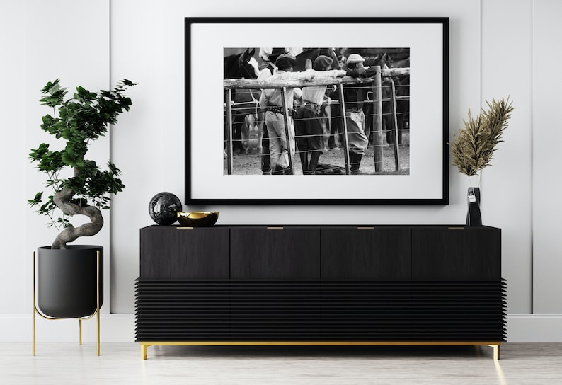 May include: Black and white framed photograph depicting gauchos leaning on a fence, observing horses. The photograph is displayed above a black wooden cabinet with gold accents, a bonsai tree, and decorative items.