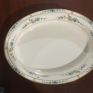 May include: An oval, off-white serving platter with a floral border. The border features a gold trim with small, colorful floral and leaf designs in blue, yellow, and red. The platter is empty and appears to be made of ceramic.