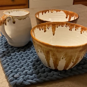 May include: A white ceramic mug and two matching bowls featuring a brown drip glaze pattern along the rims. The pieces are arranged on a blue chunky knit mat. The mug has a curved handle, and the bowls have a deep, rounded shape.