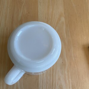 Puede incluir: Una taza de cer&aacute;mica blanca con asa, vista desde abajo. La taza est&aacute; sobre una superficie de madera marr&oacute;n claro. Un objeto rectangular negro est&aacute; en el fondo.