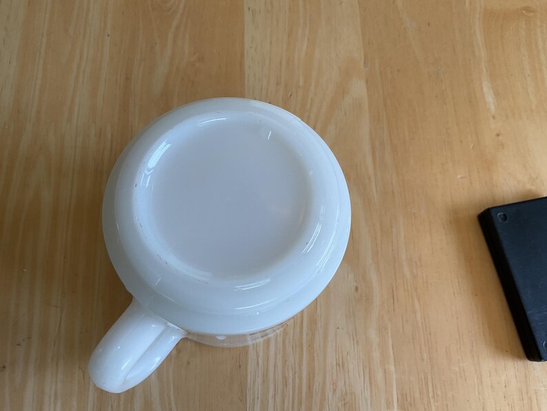 Puede incluir: Una taza de cer&aacute;mica blanca con asa, vista desde abajo. La taza est&aacute; sobre una superficie de madera marr&oacute;n claro. Un objeto rectangular negro est&aacute; en el fondo.