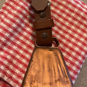 May include: A copper-colored metal bell with a trapezoidal shape attached to a dark brown leather strap. The strap features two circular metal rivets and a leather loop. The bell has a weathered, textured surface with visible vertical scratches.