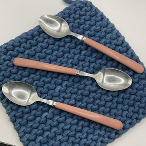 May include: Three silver spoons with pink handles rest on a blue knitted placemat. The spoons have a classic design with a rounded bowl and a smooth, solid pink handle. The placemat has a textured, woven appearance.