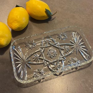 May include: A rectangular clear glass tray featuring a pressed starburst and geometric pattern with a scalloped edge, positioned next to three bright yellow lemons. The glass dish has a faceted surface that reflects light through its intricate design.