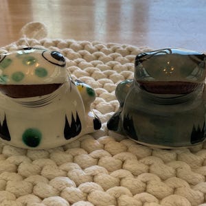 May include: Two ceramic frog figurines. One is white with green spots and black accents, the other is a solid green. Both have brown mouths and are sitting on a cream-colored, woven mat. Decorative home decor.