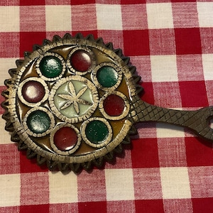 May include: Ornate, round, wooden hand mirror with a decorative design. The mirror features a central star design surrounded by red and green circular accents. The handle is textured and has a hole at the end.