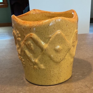 May include: A mustard yellow ceramic planter with a speckled finish and a wavy, scalloped rim. The exterior features a raised, embossed diamond pattern around the center, with additional floral-like relief details visible on the side surface.