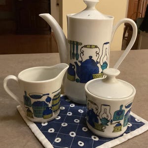 May include: A vintage white ceramic coffee set with blue and green kitchen-themed illustrations. The set includes a tall coffee pot, a creamer, and a lidded sugar bowl. The set is placed on a blue and white patterned placemat.