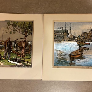 May include: Two framed vintage prints. The left print depicts a man standing near a wooden structure by the water. The right print shows a harbor scene with boats and buildings. Both prints have a watercolor style.