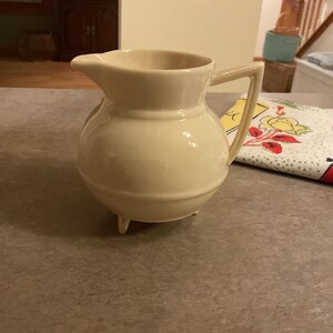 May include: A cream-colored ceramic pitcher with a rounded body, a spout, and a rectangular handle. The pitcher has three small feet and is likely vintage. The pitcher is sitting on a gray surface.