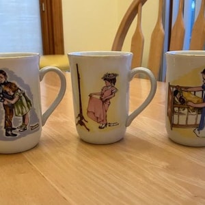 May include: Three white ceramic mugs with illustrations. One mug depicts a boy and girl with a dog, another shows a woman in a pink dress, and the last features a girl with a baby in a crib. Each mug has a curved handle.