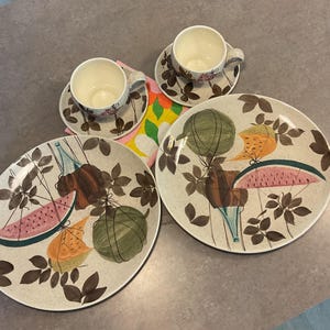 May include: A set of two ceramic dinner plates and two matching teacups with saucers, featuring a speckled cream base with brown leaf patterns and illustrations of sliced watermelon and cantaloupe suspended by thin black lines.