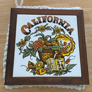 May include: A square, ceramic trivet with a wooden frame and a crocheted border. The trivet features the word "CALIFORNIA" in brown letters, along with illustrations of fruit, flowers, and a rooster.