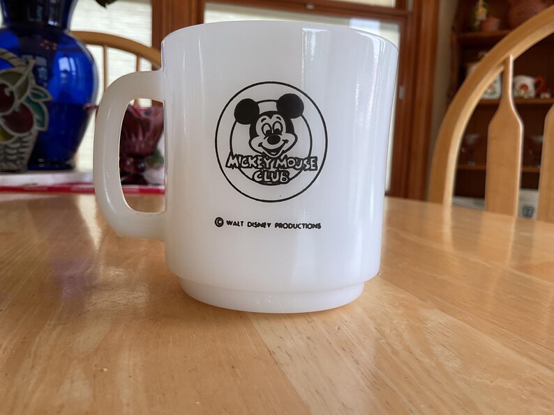 Puede incluir: Taza de cer&aacute;mica blanca con el logotipo de Mickey Mouse Club en negro. La taza tiene un asa redondeada y una base apilada. El texto "Walt Disney Productions" est&aacute; impreso debajo del logotipo. Un objeto de colecci&oacute;n vintage.