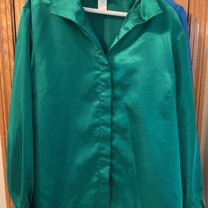 May include: A long-sleeved button-down shirt in a vibrant emerald green satin fabric. The shirt features a classic pointed collar, a concealed front button placket, and cuffs at the wrists. A white brand label is visible at the inner neckline.