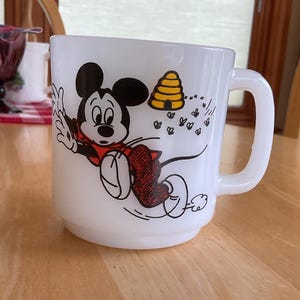 Puede incluir: Taza de cer&aacute;mica blanca con un dise&ntilde;o de dibujos animados de Mickey Mouse con un atuendo rojo, huyendo de una colmena amarilla y abejas. La taza tiene un asa curva y est&aacute; sobre una superficie de madera.