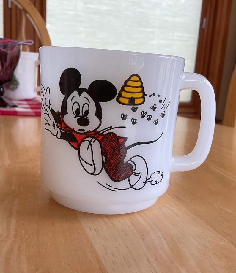 Puede incluir: Taza de cer&aacute;mica blanca con un dise&ntilde;o de dibujos animados de Mickey Mouse con un atuendo rojo, huyendo de una colmena amarilla y abejas. La taza tiene un asa curva y est&aacute; sobre una superficie de madera.
