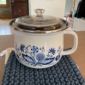 May include: White enamelware pot with a clear glass lid and a silver rim. The pot features a blue floral design around the base and a large handle. It sits on a blue knitted trivet. A vintage kitchenware item.