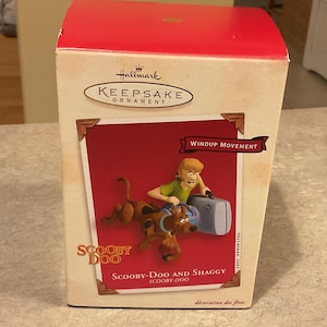 May include: A Hallmark Keepsake Ornament box featuring Scooby-Doo and Shaggy. The box displays the text Windup Movement, Scooby-Doo, and Scooby-Doo and Shaggy. The graphic shows Shaggy holding a gray suitcase while Scooby-Doo runs alongside.