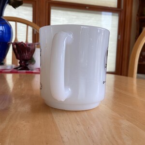 Puede incluir: Una taza de cer&aacute;mica blanca con un asa curva. La taza tiene una base ligeramente ensanchada y un acabado liso y brillante. Hay peque&ntilde;as ilustraciones negras en el lateral de la taza. La taza est&aacute; sobre una mesa de madera marr&oacute;n claro.