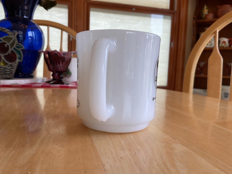 Puede incluir: Una taza de cer&aacute;mica blanca con un asa curva. La taza tiene una base ligeramente ensanchada y un acabado liso y brillante. Hay peque&ntilde;as ilustraciones negras en el lateral de la taza. La taza est&aacute; sobre una mesa de madera marr&oacute;n claro.