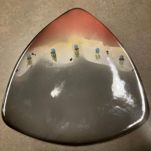 May include: Triangular ceramic plate with a gradient design. The top section transitions from red to yellow, while the lower portion is gray. Small, stylized blue and green painted details are arranged across the plate's surface.