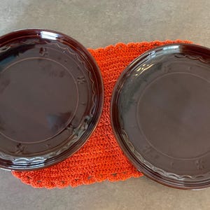 May include: Two dark brown ceramic plates with a scalloped edge and embossed floral design, resting on a bright orange crocheted mat. The plates have a glossy finish and are set against a neutral background.