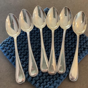 May include: Six silver spoons with oval bowls and beaded edges are arranged on a textured blue surface. The spoons are reflective, and the handles taper to a rounded end. The spoons are likely for serving or eating.